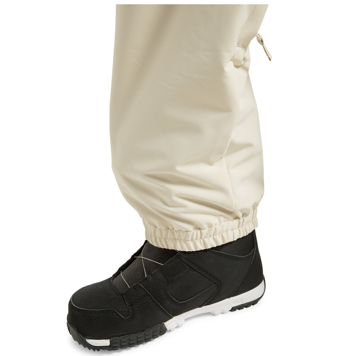 A close-up of a person wearing black athletic shoes and the DC Shoes Unisex Primo Snow Pants in cream, featuring elastic cuffs and made from recycled polyester, covering the ankle, against a white background.