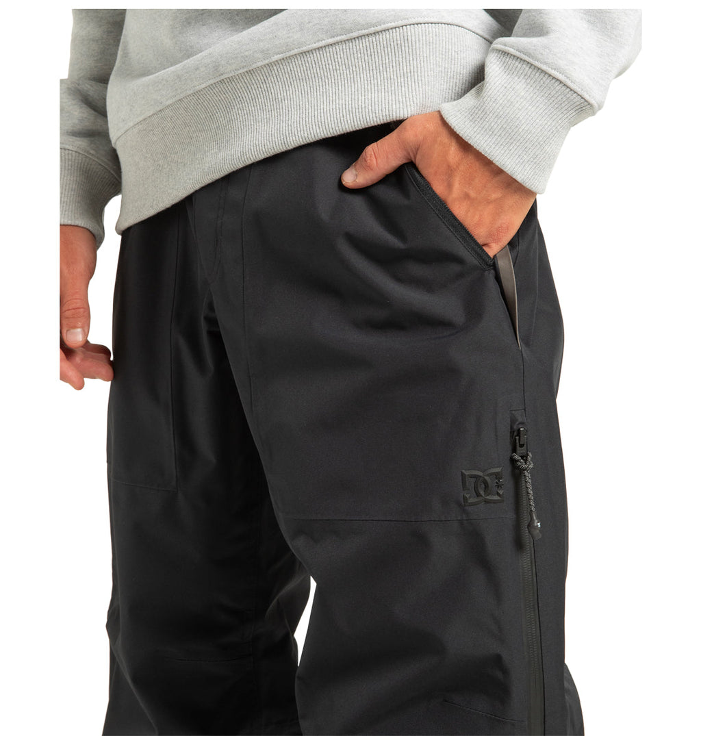 A person wearing a light gray sweatshirt and black DC Shoes Unisex Squadron 30K Insulated Pants, which feature a zipper pocket and the DC Shoes logo on the pant leg, stands with one hand in their pants pocket—these pants are ideal for winter adventures and weather defense.