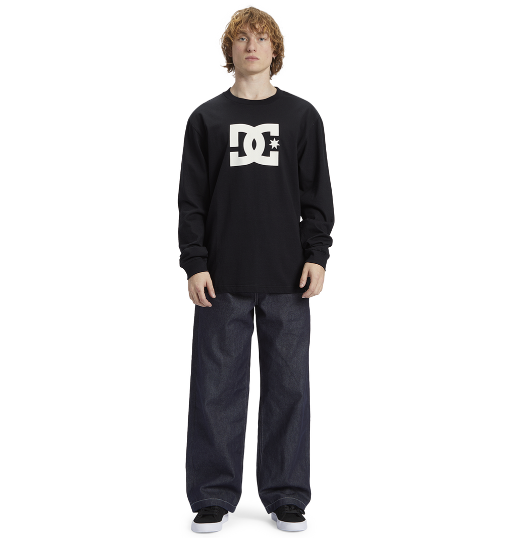 Young person with curly hair wears a black Men's DC Star Tee by DC Shoes, featuring bold graphics, paired with loose dark jeans and black sneakers, standing against a white background—ready to skate.
