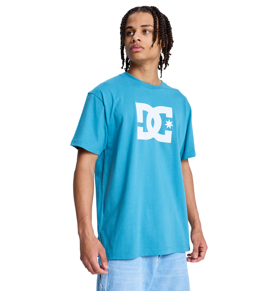 A person with braided hair wearing a bright blue Reebok DC Star Hss t-shirt and light blue jeans stands turned slightly to the side against a white background.