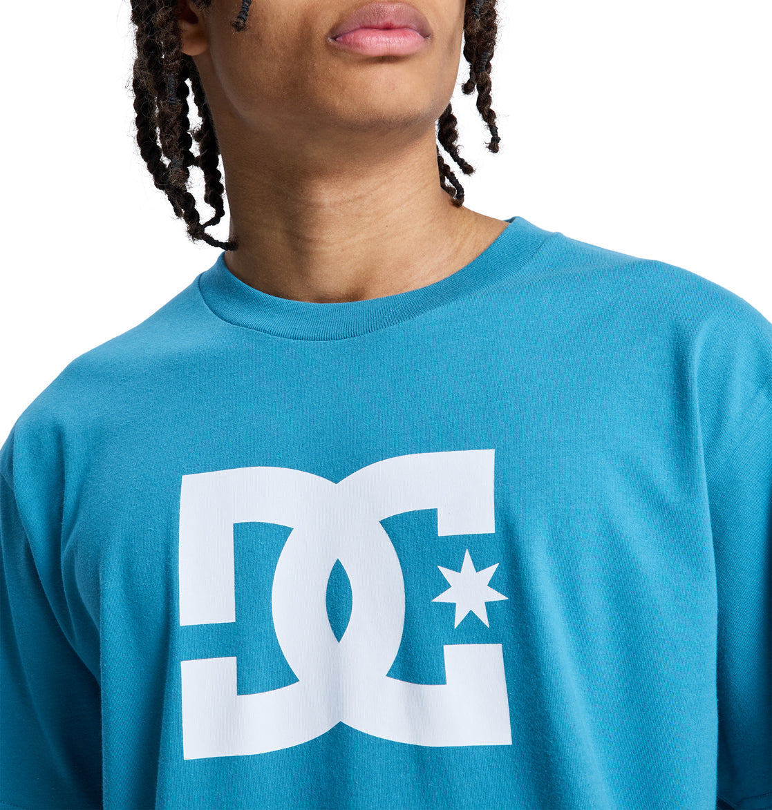 A person with braided hair wears a teal Reebok DC Star Hss T-shirt with a large white logo on the front, showing only their lower face and upper body.