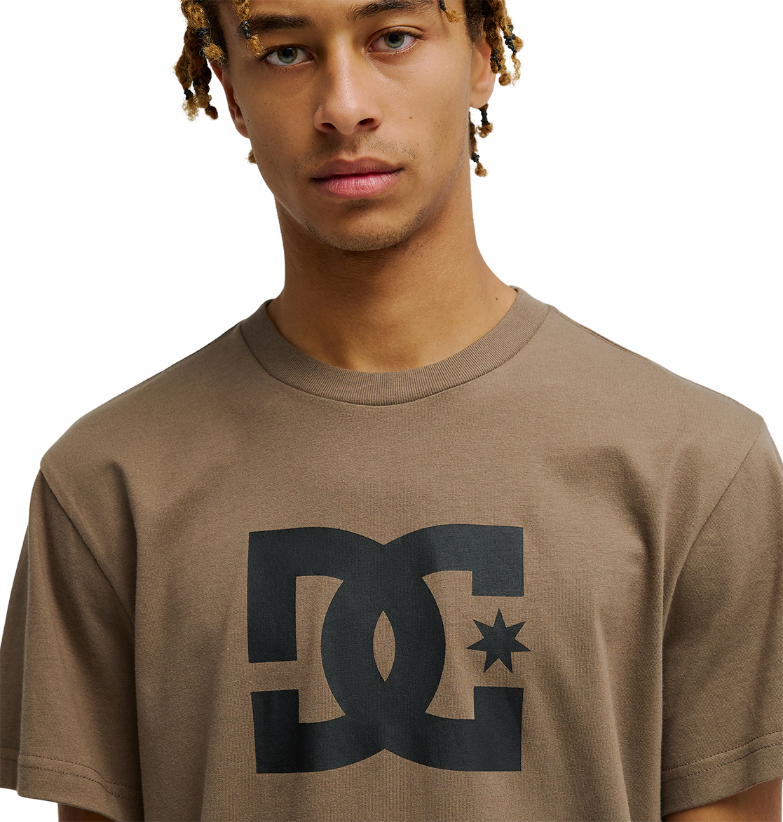 A young person with light brown skin and blonde-tipped dreadlocks wears the men's DC Star Tee by DC Shoes, a relaxed fit brown skate shirt with a bold black DC Shoes graphic on the chest, standing against a plain white background.