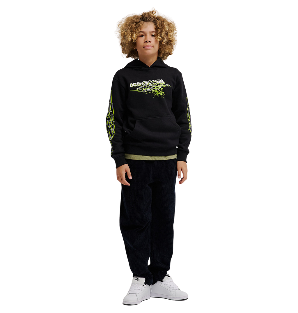A child with curly hair wears the DC Shoes Side Flame Hoodie Youth, a black fleece hoodie with neon green DC graphics, paired with dark pants and white sneakers, standing facing forward against a plain white background.