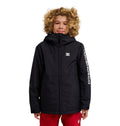 A young person with curly blond hair stands against a white background, wearing the DC Shoes Basis Youth Jacket—a black, durable, and stylish hooded jacket with DCSHOECO printed on the left sleeve—paired with red pants.