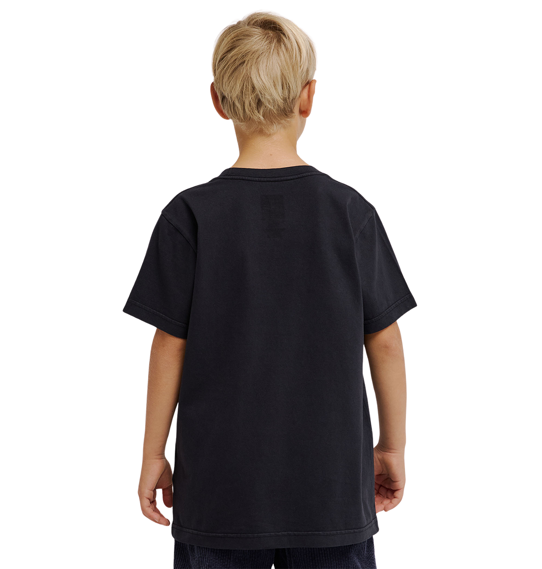 A young boy with short blond hair is standing with his back to the camera, wearing a loose-fitting, plain black DC Speratic Tee Youth by DC Shoes and dark shorts against a light gray background.