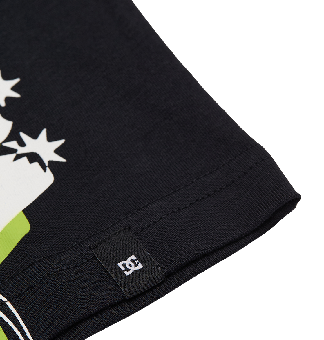 Close-up of the black fabric edge of the DC Side Flame Tee Youth by DC Shoes, featuring a small stitched tag with a white DC logo. Partially visible on the fabric are colorful graphic elements and some white star shapes from the skate tee’s design.