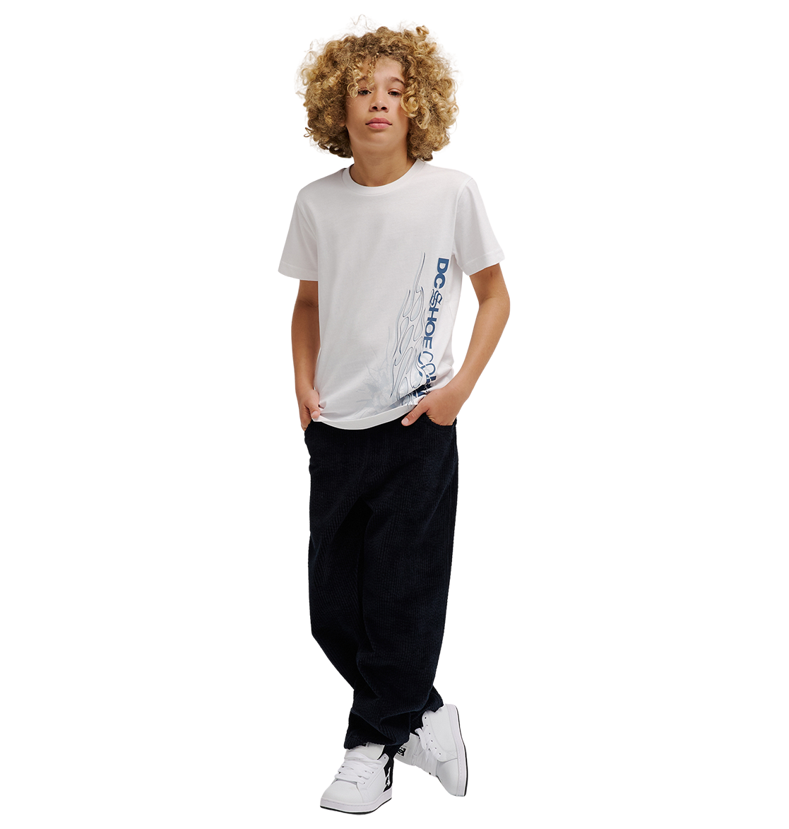 A boy with curly hair wears the DC Shoes DC Side Flame Tee Youth, a white graphic skate tee featuring bold flame artwork, paired with black relaxed fit pants and white sneakers. He stands with his hands in his pockets against a plain white background.