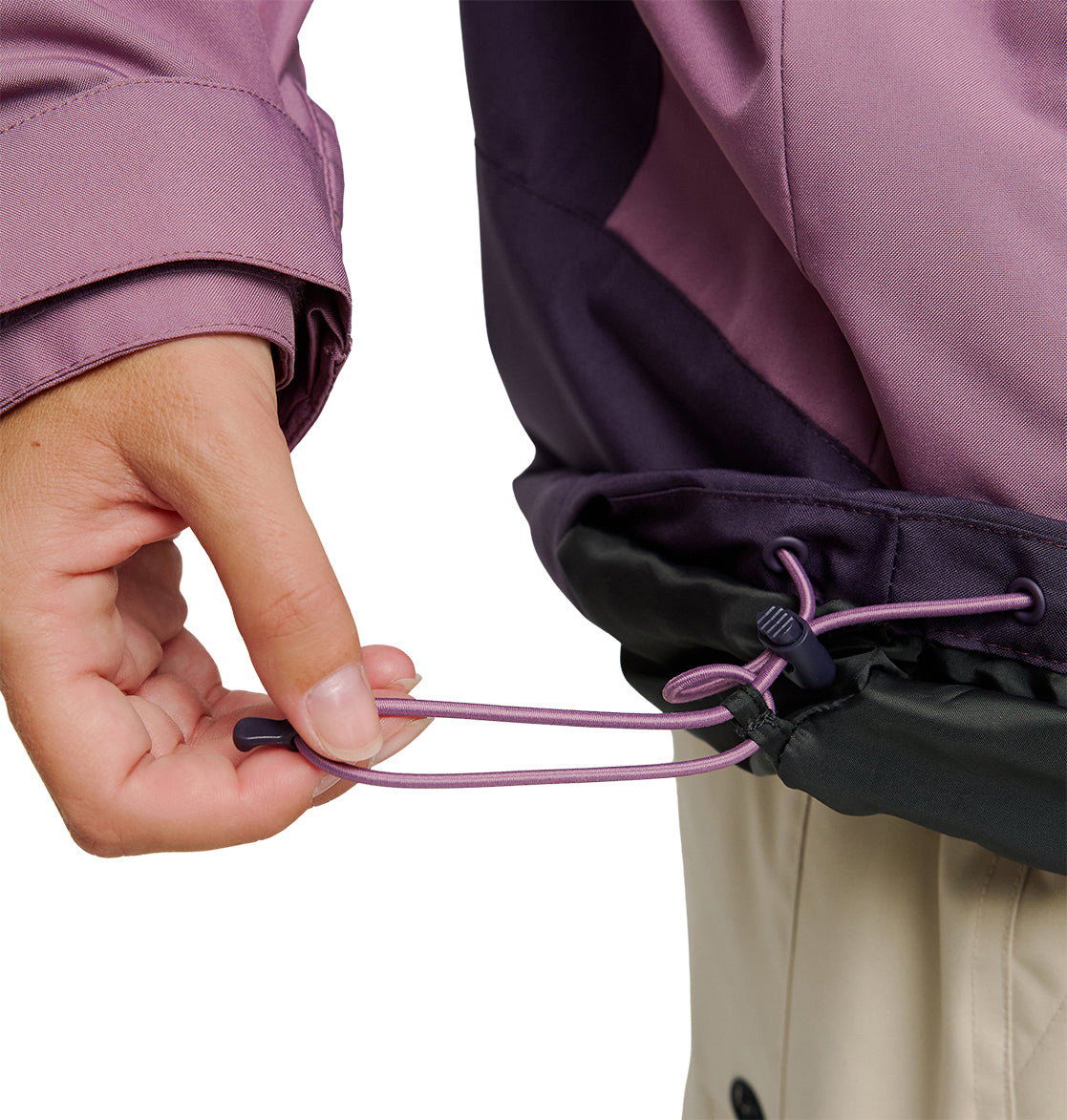 A hand pulls a purple drawstring at the hem of the DC Shoes Everfrost Jacket—a two-tone purple and black insulated style—adjusting its fit. The person is wearing light-colored pants.