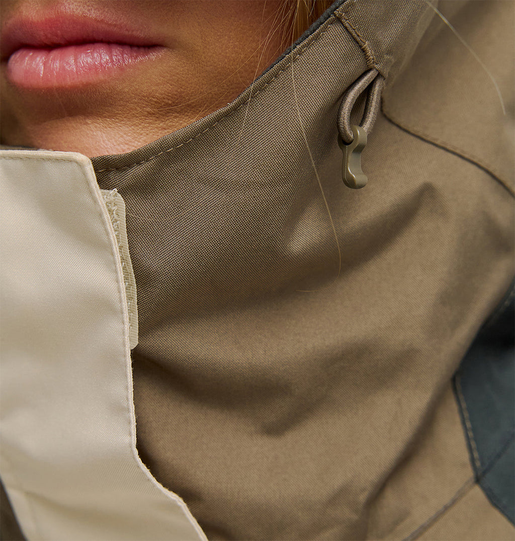 Close-up of a person’s lower face, showing pink lips and part of the chin. The person is wearing the DC Shoes Everfrost Jacket—an insulated outerwear piece in tan and beige, featuring a high collar with a visible drawstring.