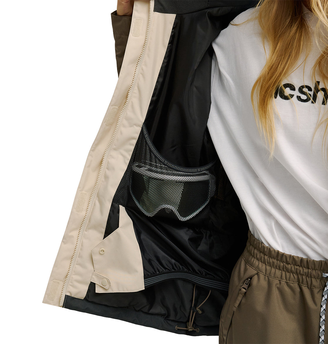 A person opens a beige DC Shoes Everfrost Jacket, revealing ski goggles stored in an inner mesh pocket. They wear a white shirt and olive green pants.