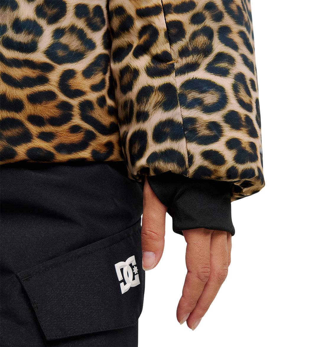 A close-up of a person’s hand by their side, wearing the DC Shoes Aura Puffer Jacket in a leopard print design and black pants with a white DC Shoes logo on the pocket.