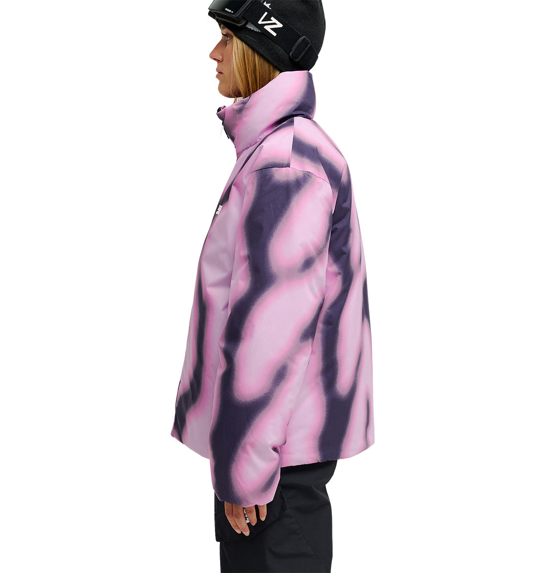 A person in a black helmet and black pants is wearing the DC Shoes Aura Puffer Jacket—a pink and purple tie-dye waterproof jacket—standing in profile against a white background.