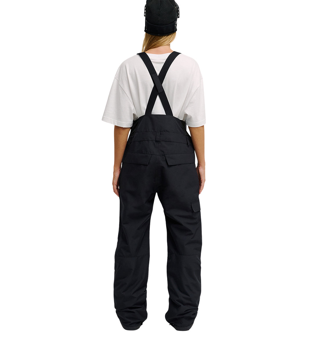 A person stands with their back to the camera against a plain white background, wearing a white t-shirt, black cap, and DC Shoes Valiant Bib Pants featuring crossed straps designed for breathable protection.