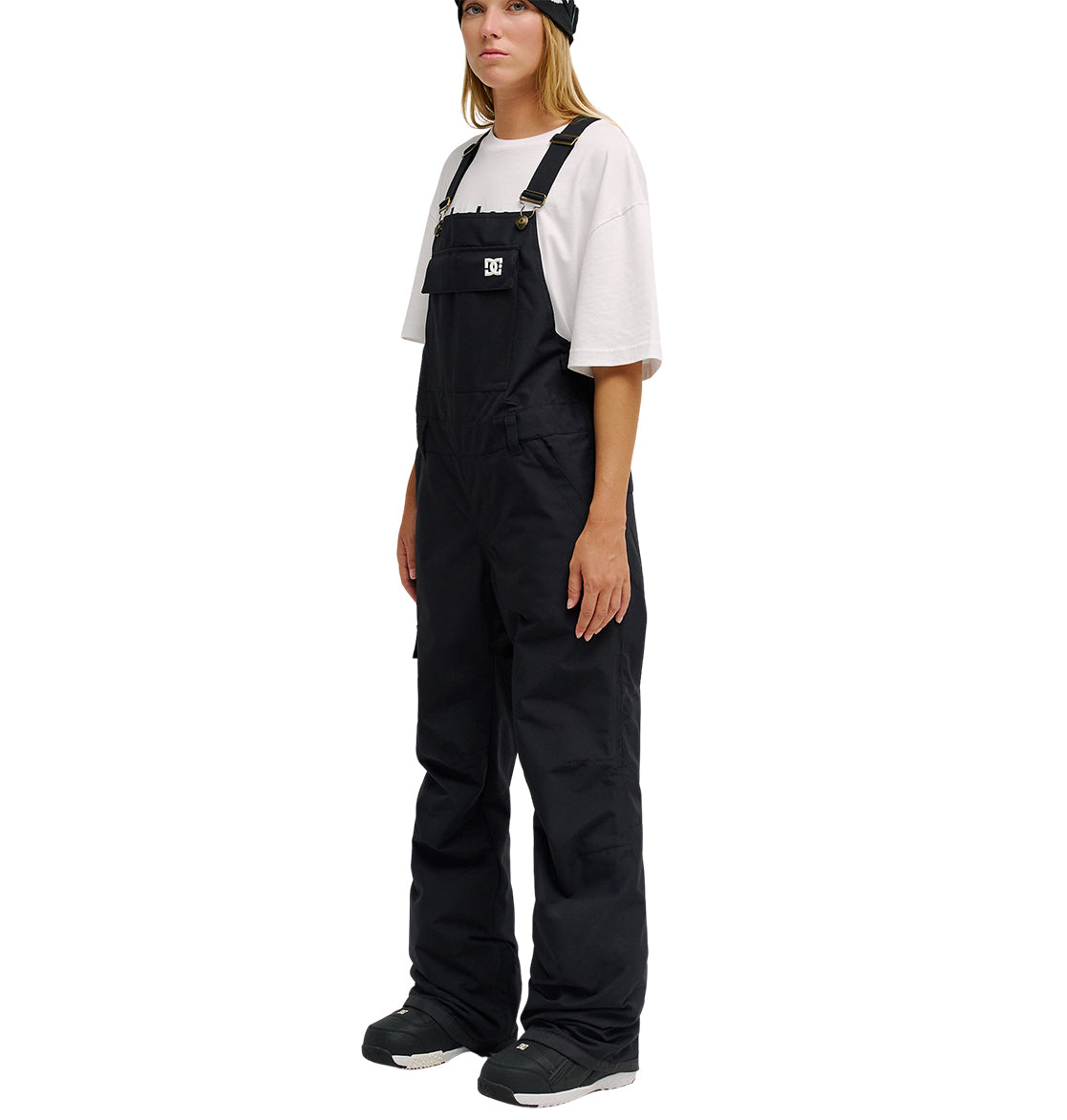 A person wearing a white t-shirt, black beanie, black boots, and DC Shoes Valiant Bib Pants stands facing slightly to the side against a white background.