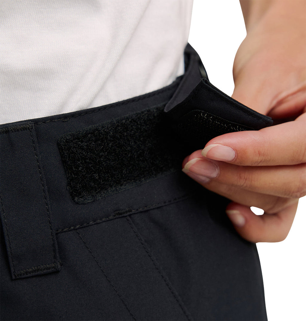 A close-up of a person fastening the Nonchalant Pant from DC Shoes with a Velcro strap, showcasing the black insulated design. The individual, dressed in a white shirt, is adjusting the strap near the waistband to emphasize features crafted for superior snow performance.