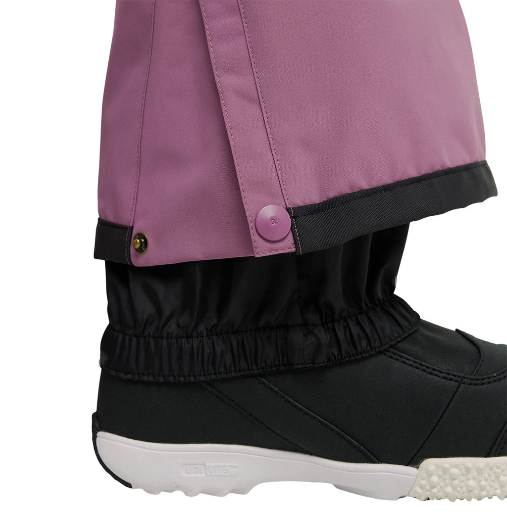 A close-up of a person’s foot wearing a black shoe and black gaiter, paired with DC Shoes Nonchalant Pants in purple. The insulated pants feature a snap button at the ankle, set against a plain white background that highlights their waterproofing and snow performance.