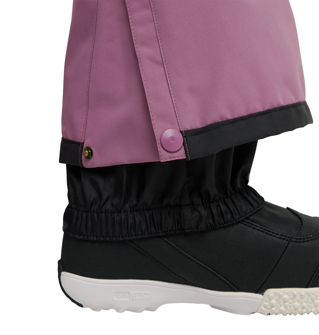 A close-up of a person’s foot wearing a black shoe and black gaiter, paired with DC Shoes Nonchalant Pants in purple. The insulated pants feature a snap button at the ankle, set against a plain white background that highlights their waterproofing and snow performance.
