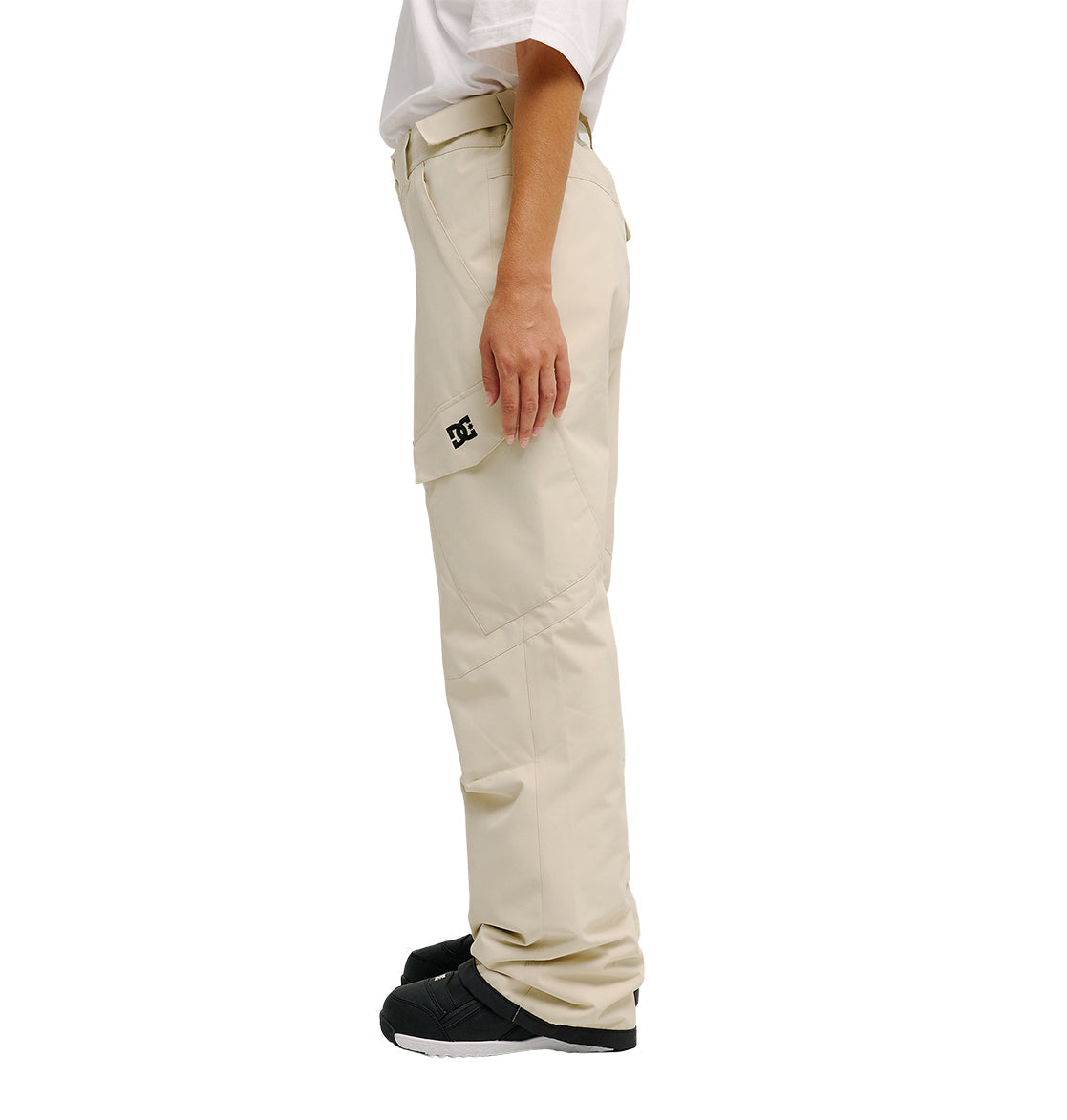 A person stands sideways against a plain white background wearing the Nonchalant Pants by DC Shoes—beige, waterproof, and insulated cargo pants featuring a side pocket with a small black DC logo—paired with a white t-shirt and black shoes.