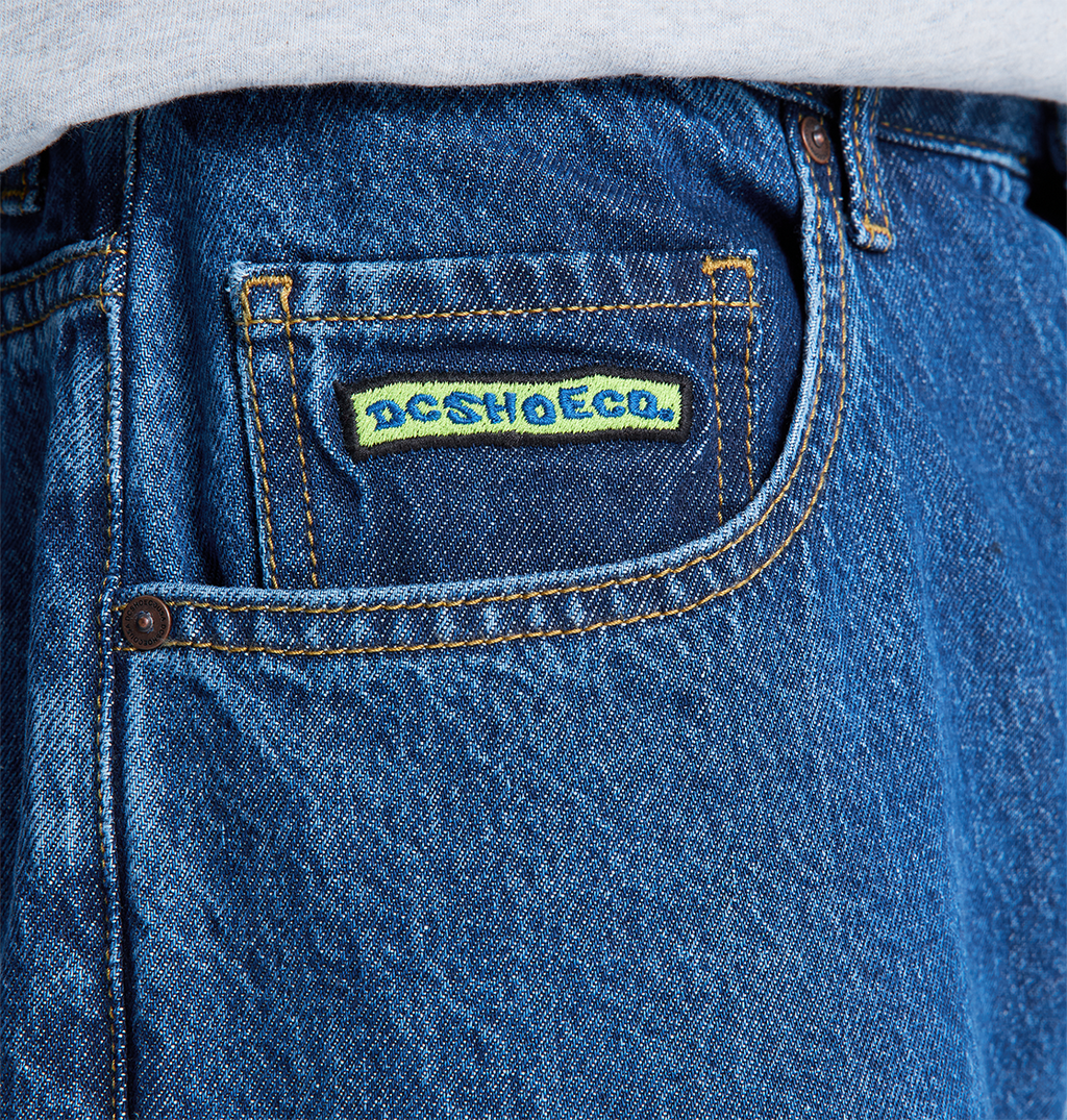 Close-up of the front pocket on DC Shoes Men's Baggy Denim Pant in blue heavyweight denim, featuring a yellow and blue “DC SHOESCO.” label above the pocket, with a white shirt partially visible at the top.