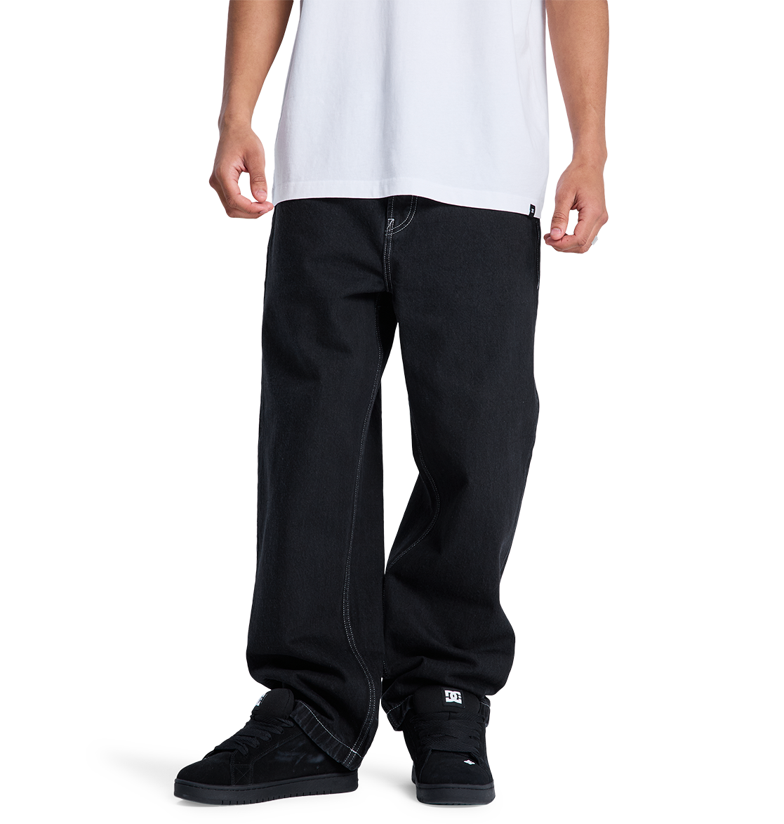 A person stands against a plain white background, wearing DC Shoes Men's Baggy Denim Pant in heavyweight denim with contrast stitching and a loose white T-shirt. The black sneakers are also visible. The image is cropped from the shoulders down.