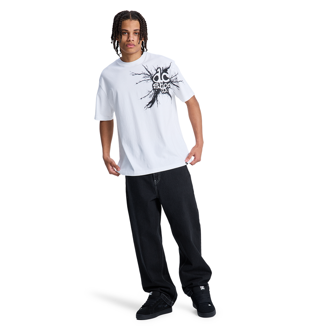 A person with braided hair wears a white graphic tee, black DC Shoes sneakers, and the Men's Baggy Denim Pant by DC Shoes, all set against a plain white background.