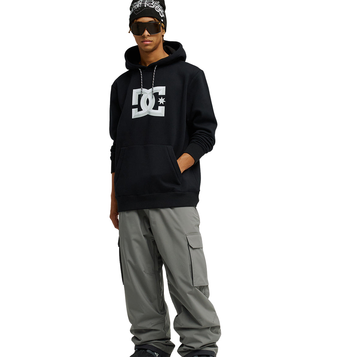 A person wearing the Snowstar black hoodie from DC Shoes with a comfortable fit, gray cargo pants, sunglasses, a black beanie, and a chain necklace stands with one hand in their pocket against a white background.