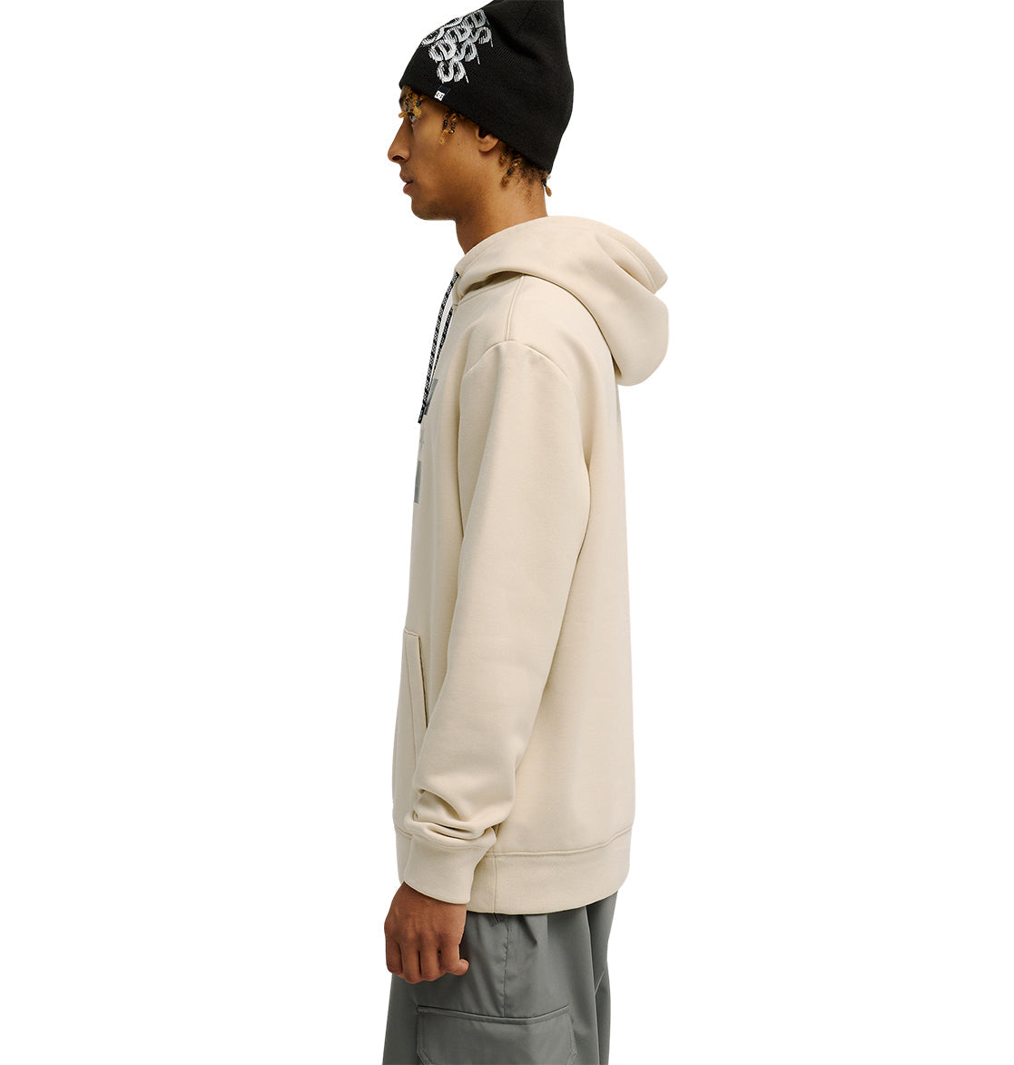 Wearing the DC Shoes Snowstar hoodie in beige, paired with gray pants and a black beanie featuring a white design, a person stands in profile against a white background, highlighting the comfortable fit and weather protection this outfit offers.