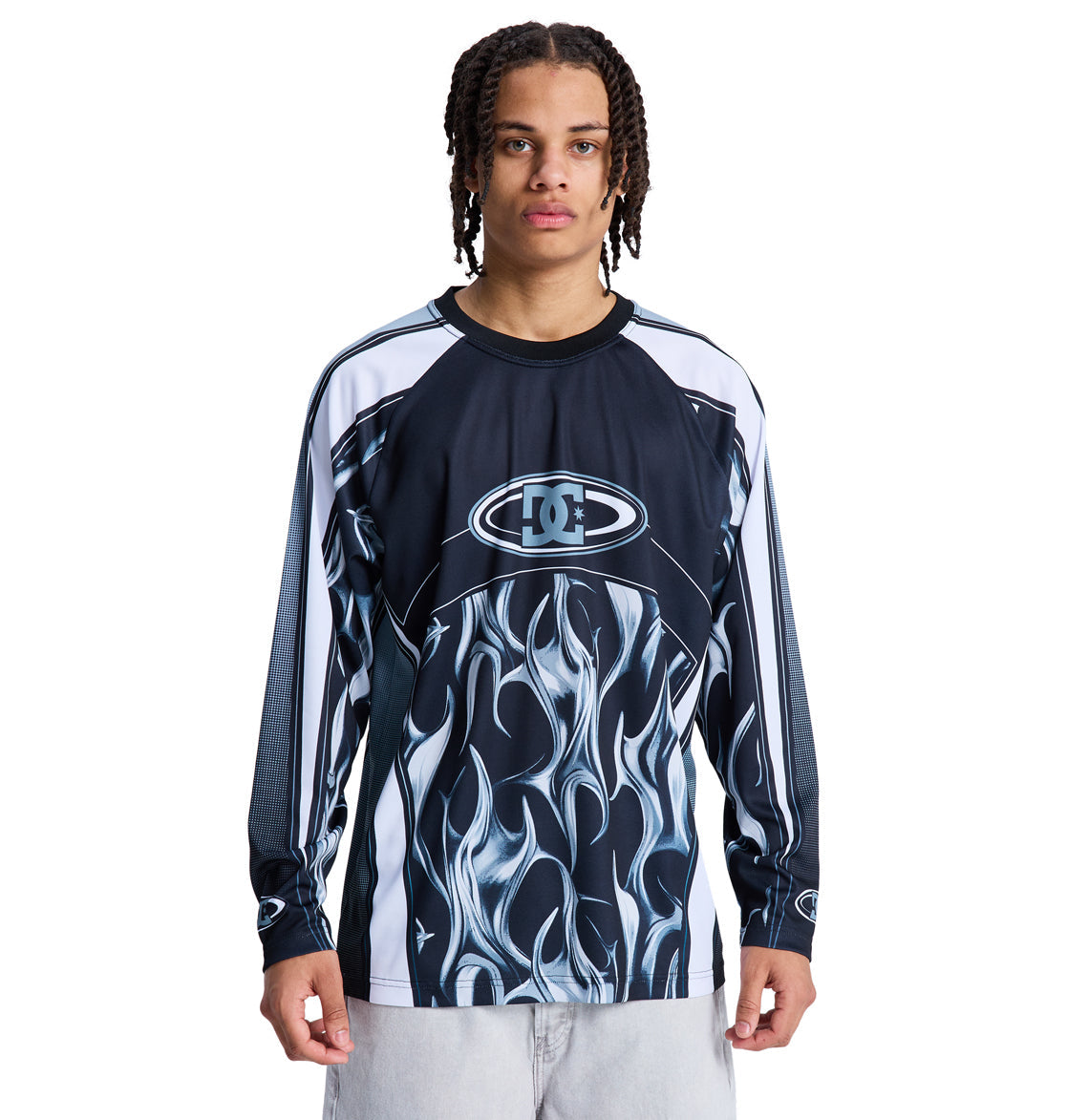 A person with braided hair wears the Reebok Sylem 2 Ls, a black and white long-sleeve shirt with a bold flame graphic, standing against a plain white background.