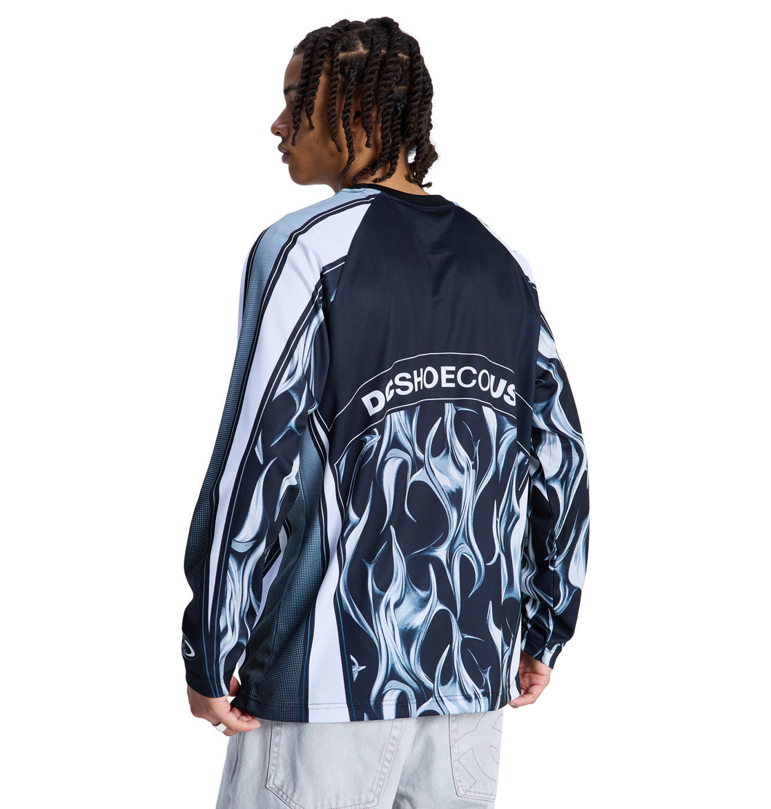 A person with braided hair wears the Reebok Sylem 2 Ls, a long-sleeve shirt with blue and white flame patterns and "DCSHOECOUSA" across the back, standing against a white background.