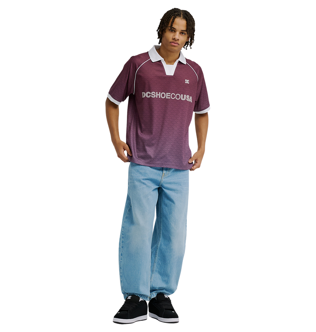A person stands against a plain white background wearing the Men's DC Soccer Top by DC Shoes in maroon, paired with light blue baggy jeans and black sneakers.