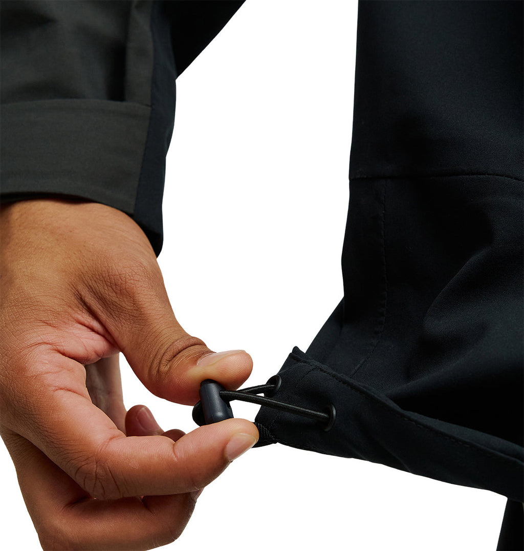 A hand adjusts the drawstring toggle on the hem of a DC Shoes Storm Division 3L Jacket 30K, tightening the fit on this black waterproof jacket for breathable protection.