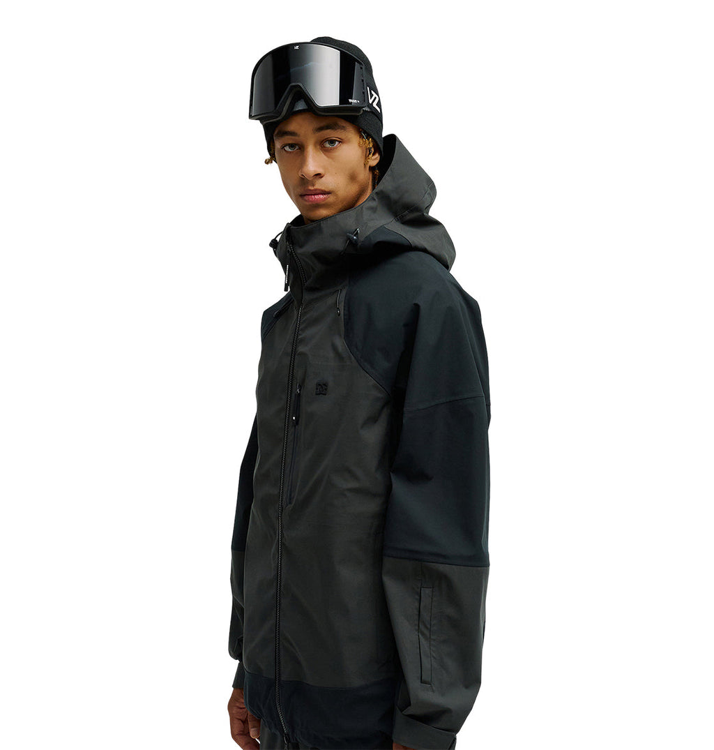 A person wearing the DC Shoes Storm Division 3L Jacket 30K in a dark color, paired with ski goggles, stands facing slightly to the side against a plain white background.