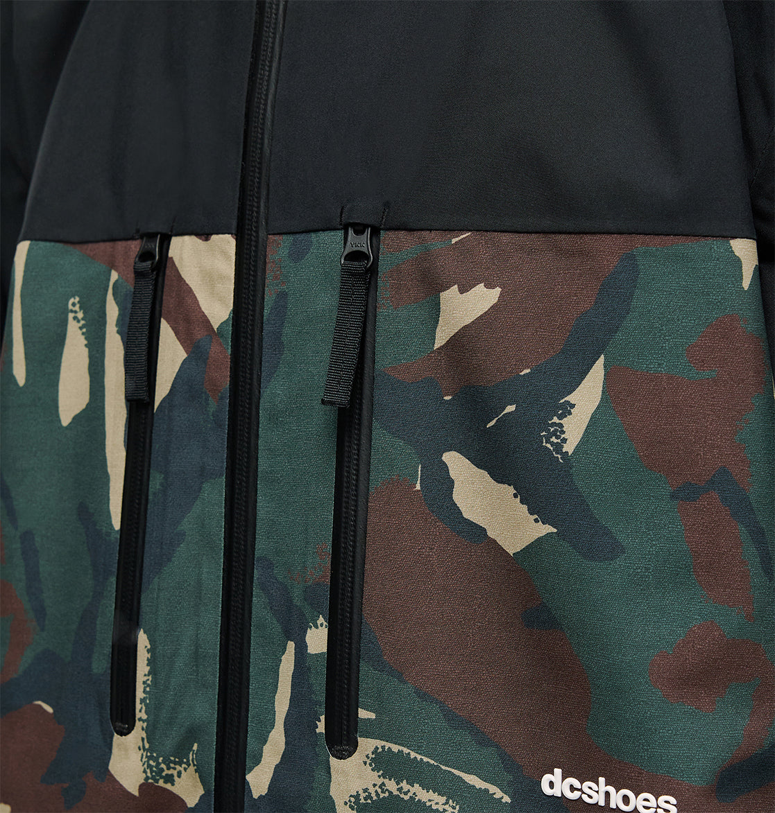 Close-up of the DC Shoes Edge Jacket 20K, highlighting its black upper fabric and camouflage pattern on the lower section. This jacket offers 20K waterproofing for superior snow performance and features two vertical zippered pockets, along with a small white DC Shoes logo on the bottom right.