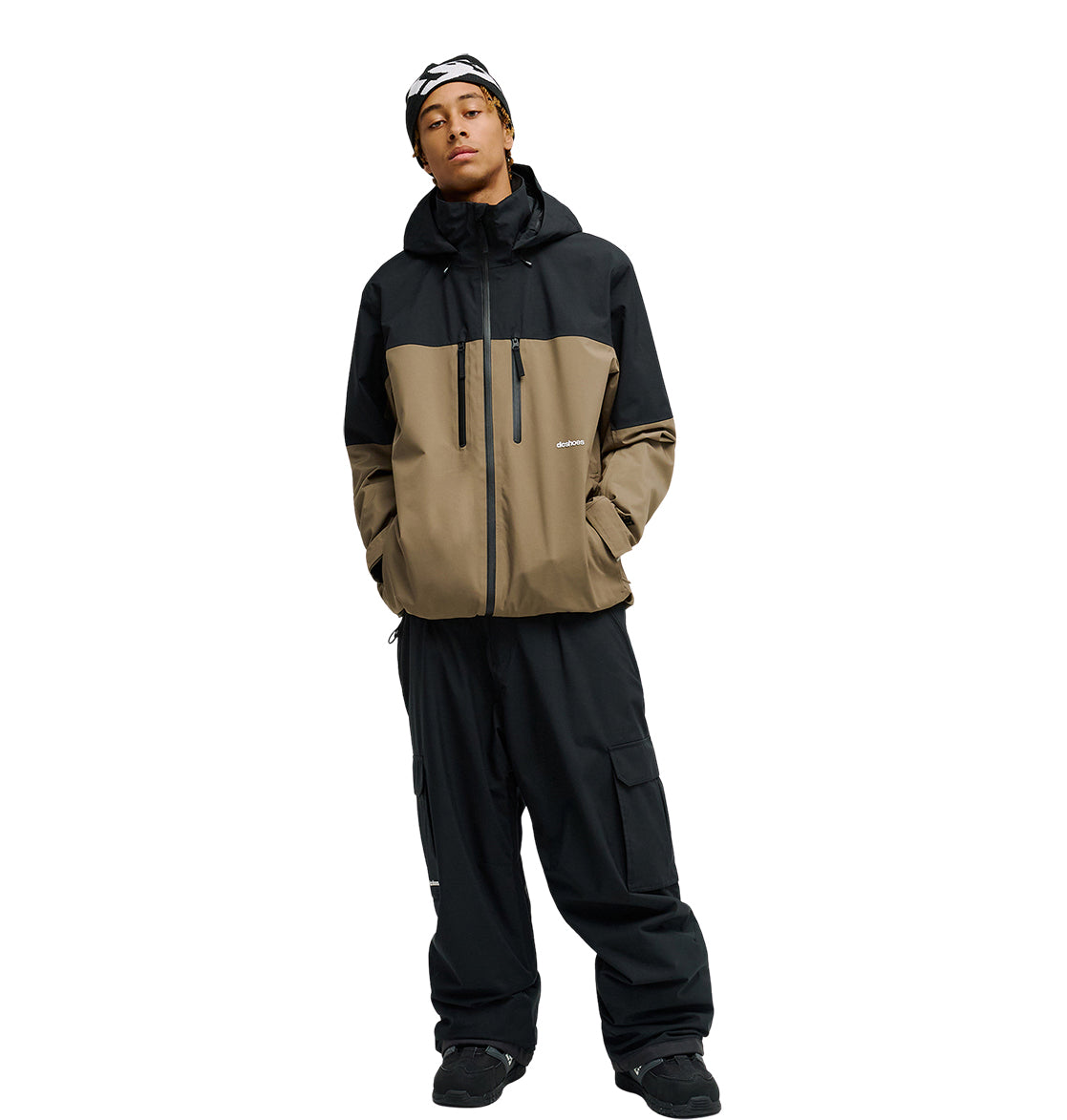A person stands against a plain white background wearing the DC Shoes Edge Jacket 20K—a two-tone black and brown waterproof jacket—paired with black cargo pants, black shoes, and a black and white beanie.