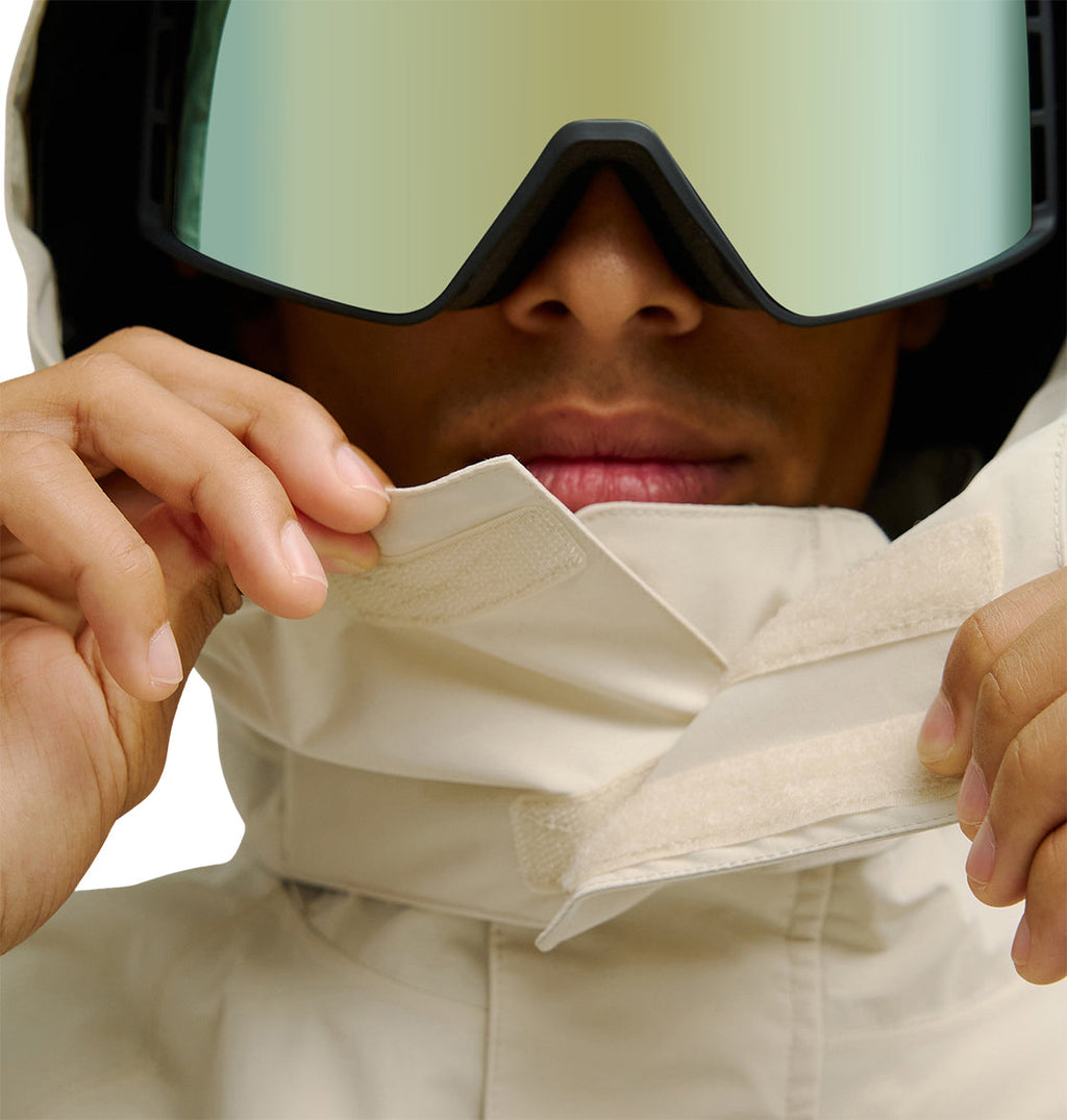 Wearing large reflective ski goggles, a person fastens the Velcro collar of the DC Shoes Mountain Lab Jacket 20K, highlighting its snow performance with only their lower face visible.