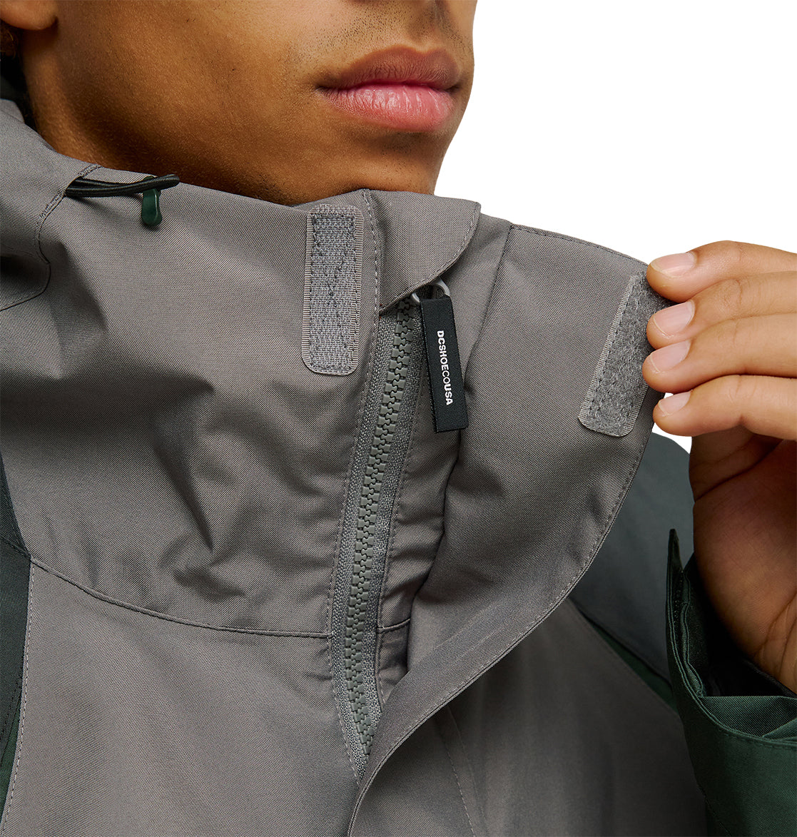 A person fastens the high collar of the gray DC Shoes Coldsmoke Jacket 10K using Velcro tabs, with part of a zipper and a small zipper pull visible.