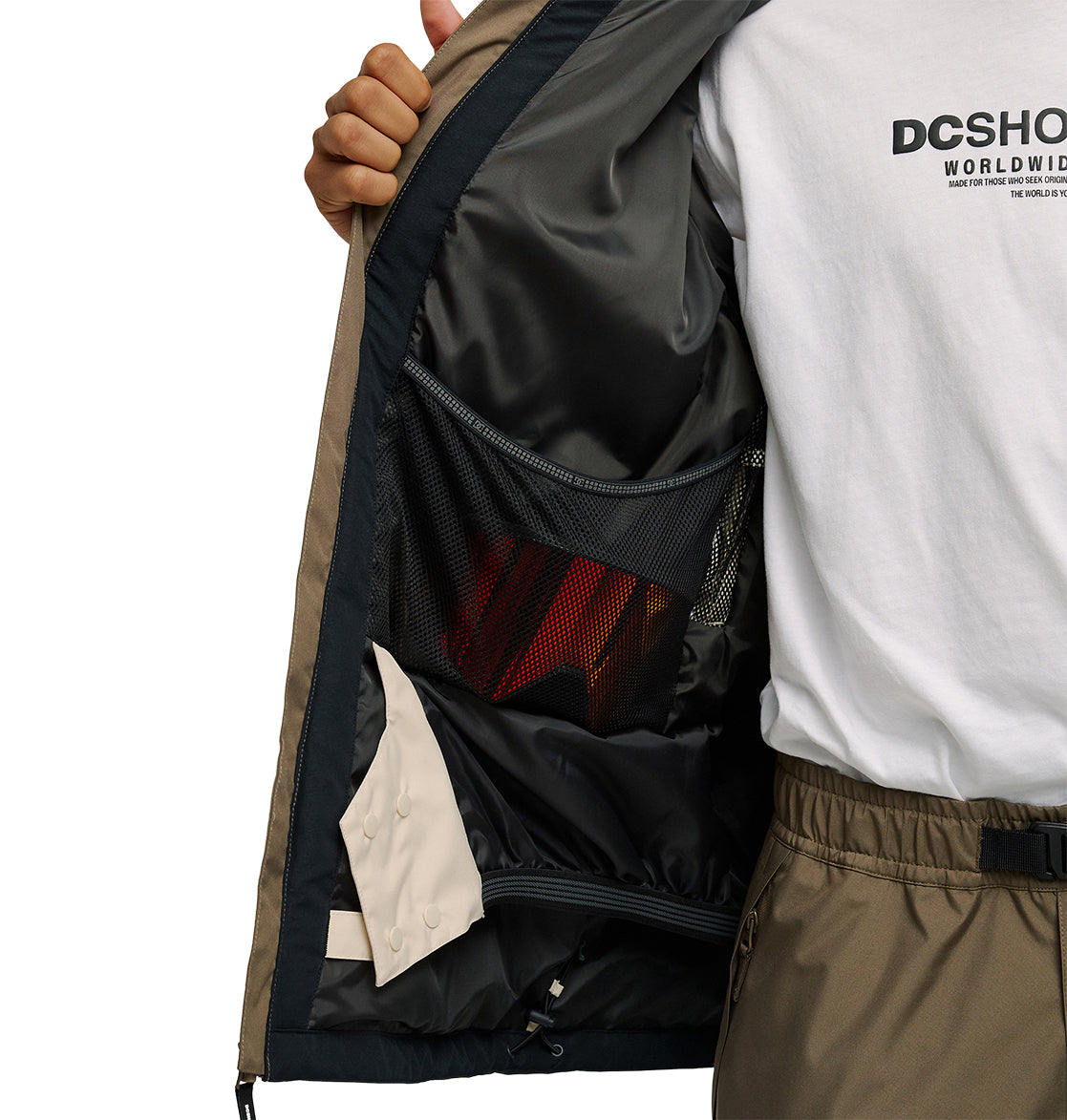 Wearing the DC Shoes Coldsmoke Jacket 10K in tan, the person opens it to reveal a mesh inner pocket holding red sunglasses. Paired with a white DC Shoes shirt and khaki pants, they effortlessly showcase style and breathable protection. Only the right side of their torso and hand are visible.