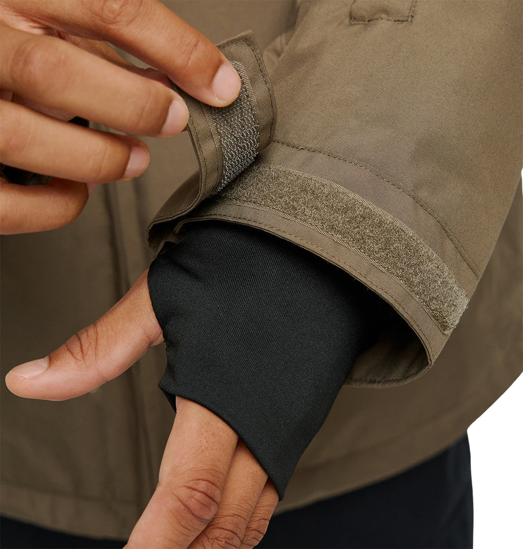 A person adjusts the Velcro cuff of a brown DC Shoes Basis Jacket 10K sleeve, revealing a black inner sleeve with a thumb hole over their hand. This insulated, breathable jacket from DC Shoes ensures comfort and protection, making it ideal for challenging conditions.