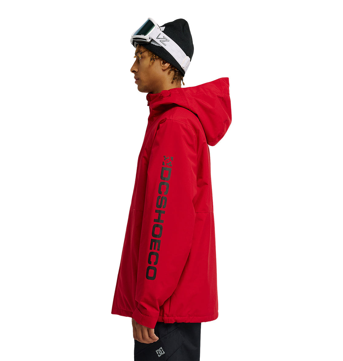 A person wearing a red DC Shoes Basis Jacket 10K with “DCSHOECO” on the sleeve, black pants, a black beanie, and white ski goggles stands in profile against a white background.