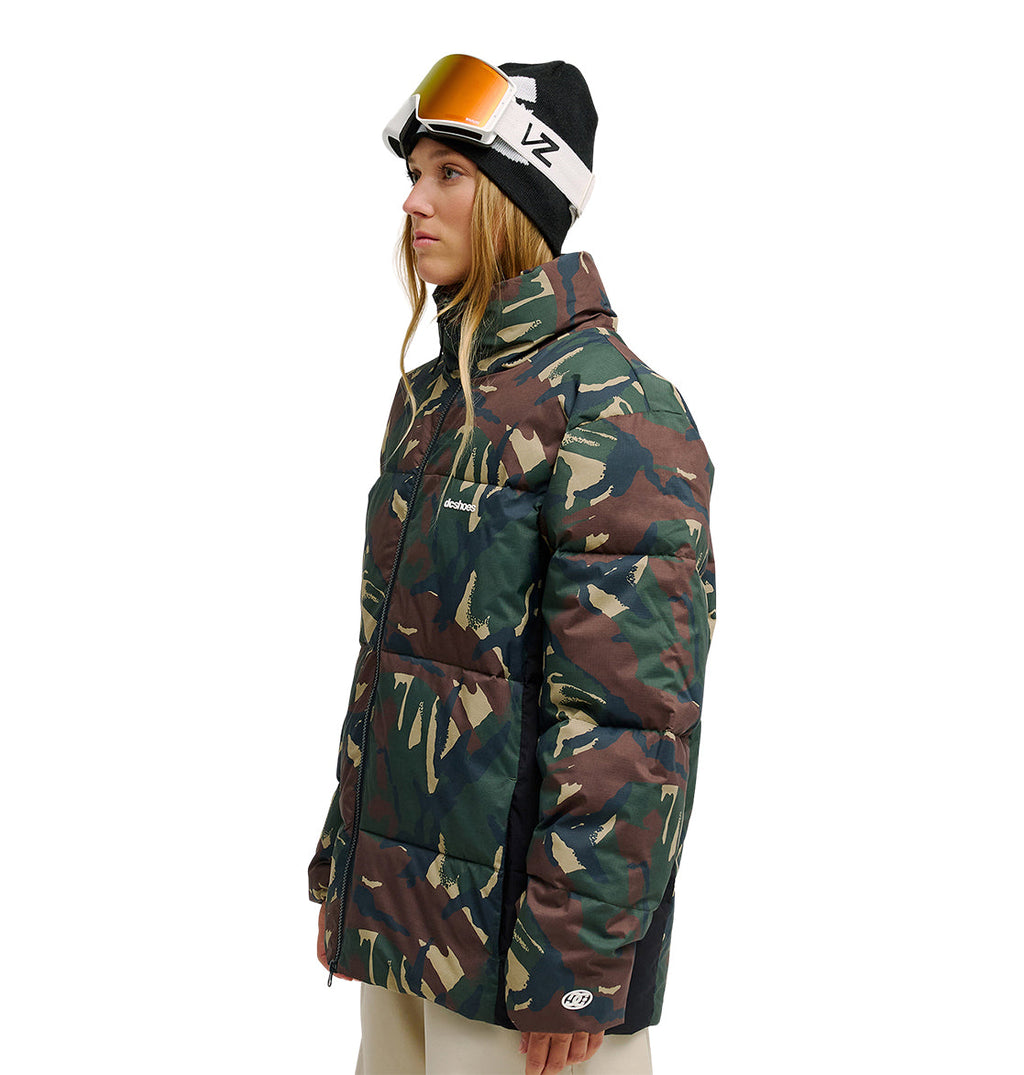 A woman stands in profile against a white background, wearing the DC Shoes Trackstar Puffer 10K insulated camouflage jacket, beige pants, and a black beanie with ski goggles.