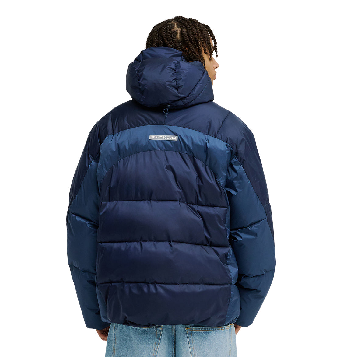 A person wearing the DC Shoes Powder Puffer 10K jacket—a blue puffer with a hood and excellent insulation—paired with light blue jeans is seen from behind. The jacket features a small rectangular label on the back, and the person’s hair is styled in short twists.