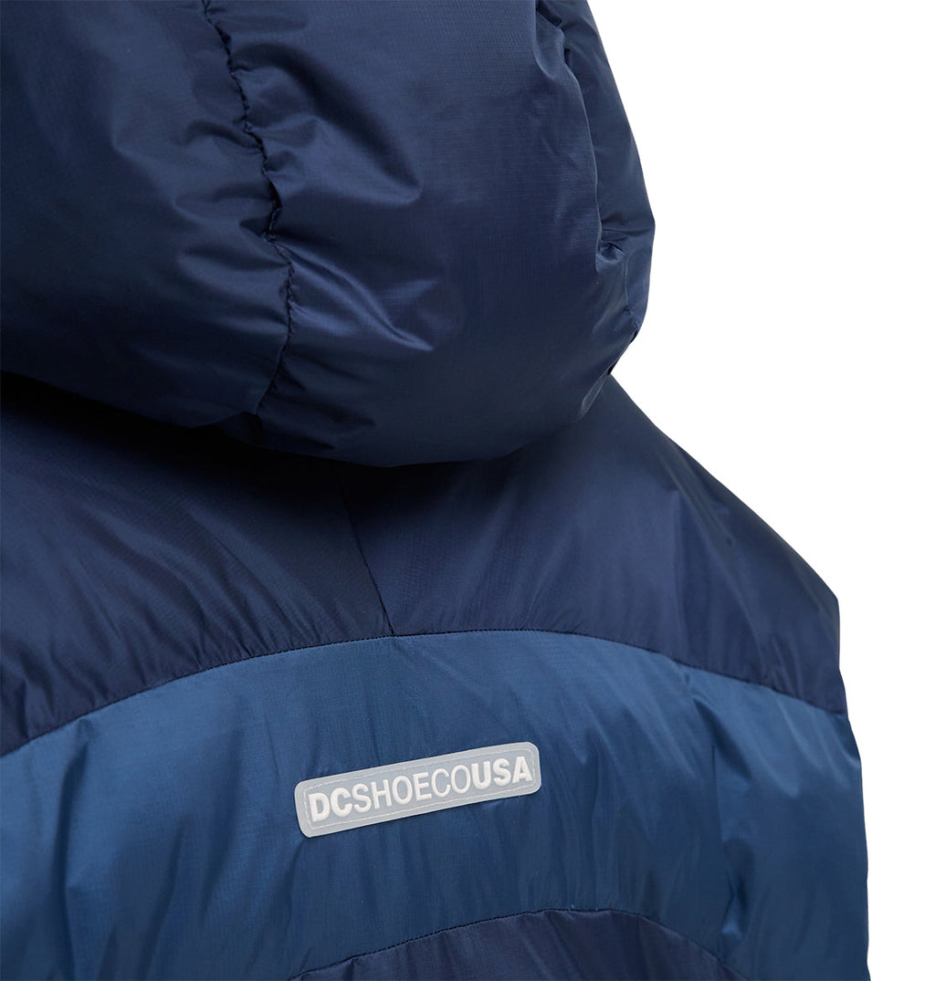 Close-up of the back of the DC Shoes Powder Puffer 10K jacket in navy blue, featuring a hood and engineered for excellent snow performance. A small rectangular label on the back displays “DCSHOECOUSA” in white text against a plain white background.