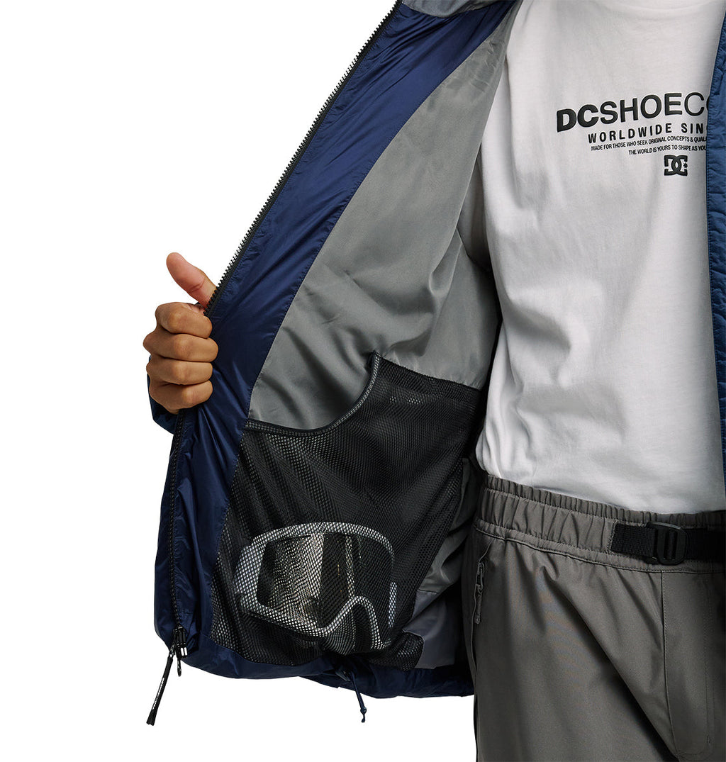 A person opens a blue DC Shoes Powder Puffer 10K jacket—crafted for superior insulation and snow performance—revealing a mesh inner pocket holding ski goggles. They wear gray pants and a white DC Shoes t-shirt.