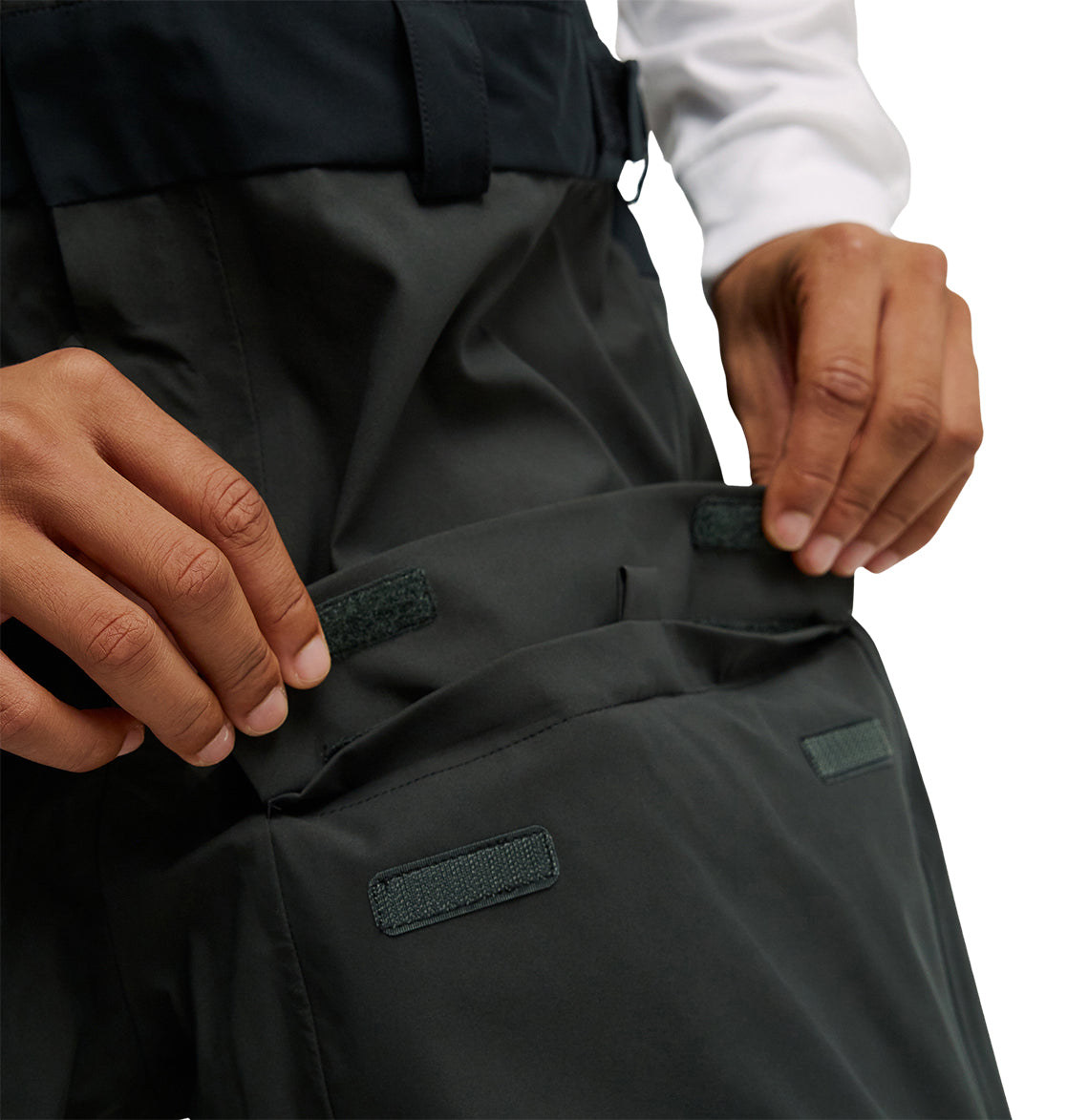 A person opens the Velcro flap of a dark-colored cargo pocket on the DC Shoes Storm Division 3L Bib 30K, showing details of the closure and highlighting the breathable protection integrated into its design.