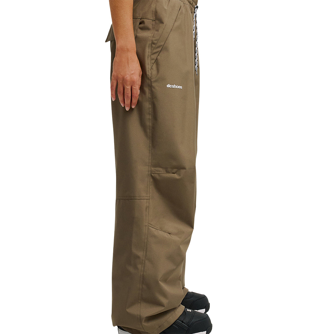 A person stands sideways wearing loose brown DC Shoes Trackstar Pants 10K with pockets and black shoes. The pants, featuring breathable waterproof protection, have the word “detchos” printed near the pocket. Only the lower half of the body is visible.