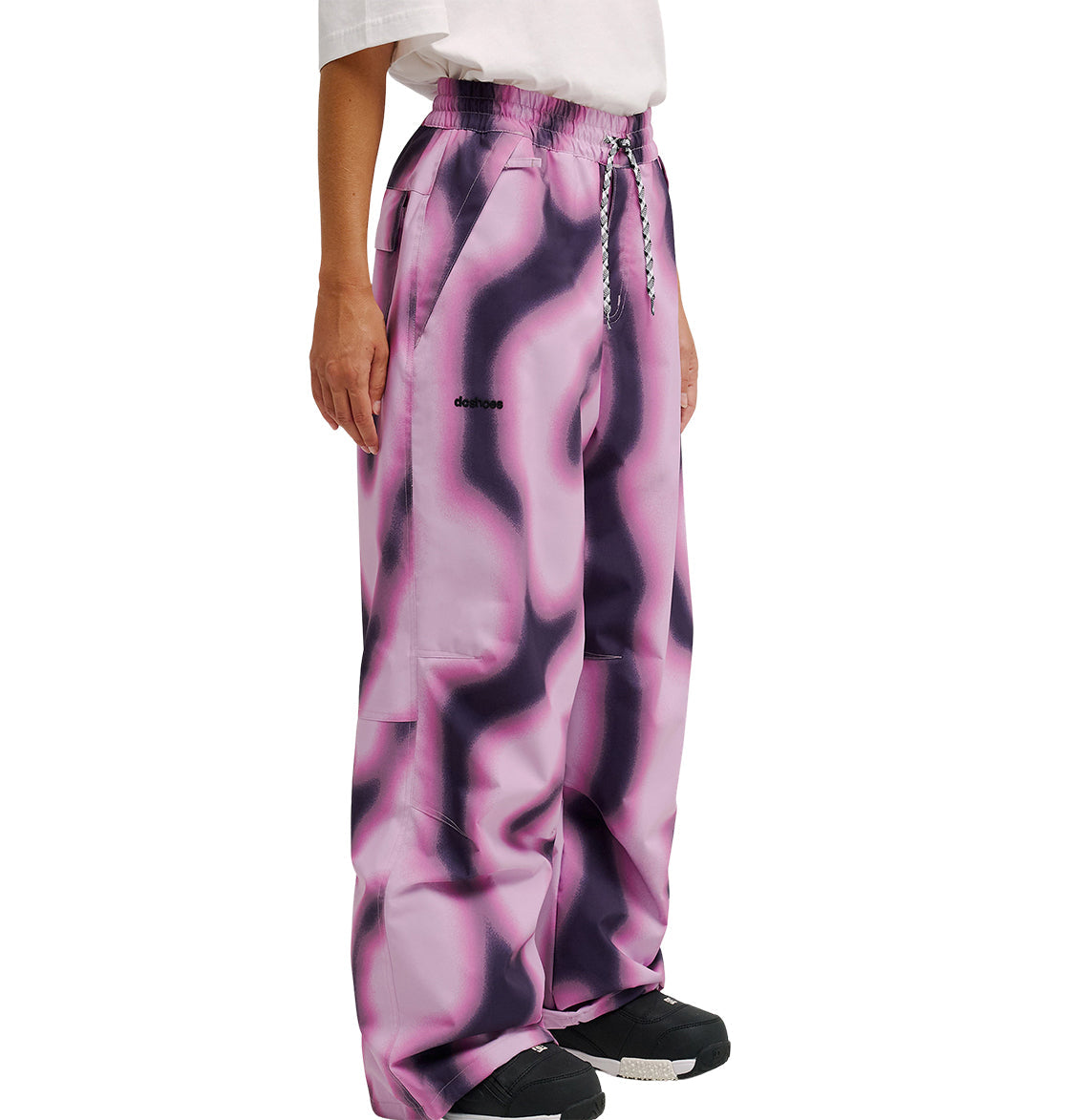 A person wearing DC Shoes Trackstar Pants 10K, featuring a loose fit and pink and black wavy pattern, paired with a white shirt and black sneakers, shown from the shoulders down against a white background.