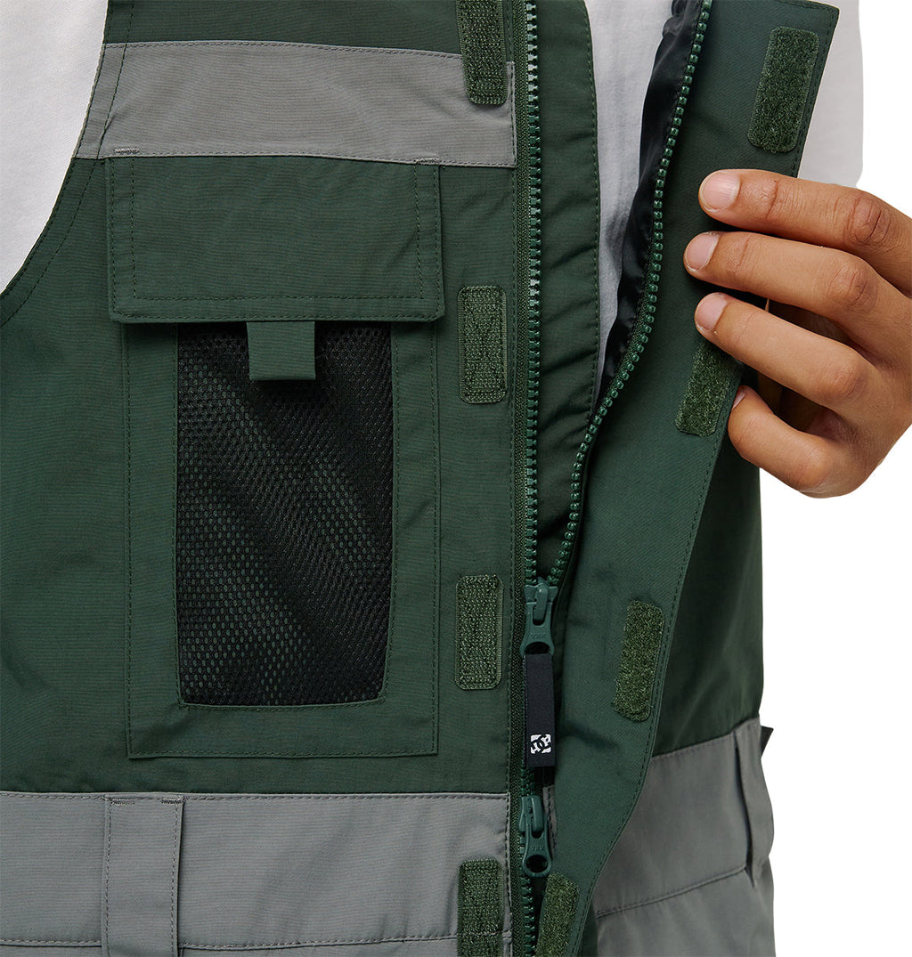 A person’s hand pulls back the front flap of a green and gray DC Shoes Mountain Lab Bib 20K, revealing a zipper and Velcro strips. Designed for breathable protection, this bib features a mesh pocket and is worn over a white shirt.