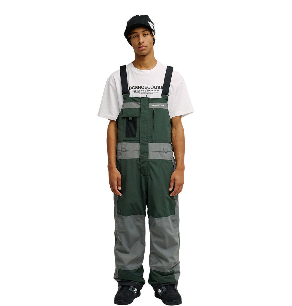 Wearing the DC Shoes Mountain Lab Bib 20K in green and gray, along with a black beanie and a white graphic T-shirt, a person stands facing forward with arms relaxed at their sides against a plain white background.