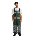 Wearing the DC Shoes Mountain Lab Bib 20K in green and gray, along with a black beanie and a white graphic T-shirt, a person stands facing forward with arms relaxed at their sides against a plain white background.