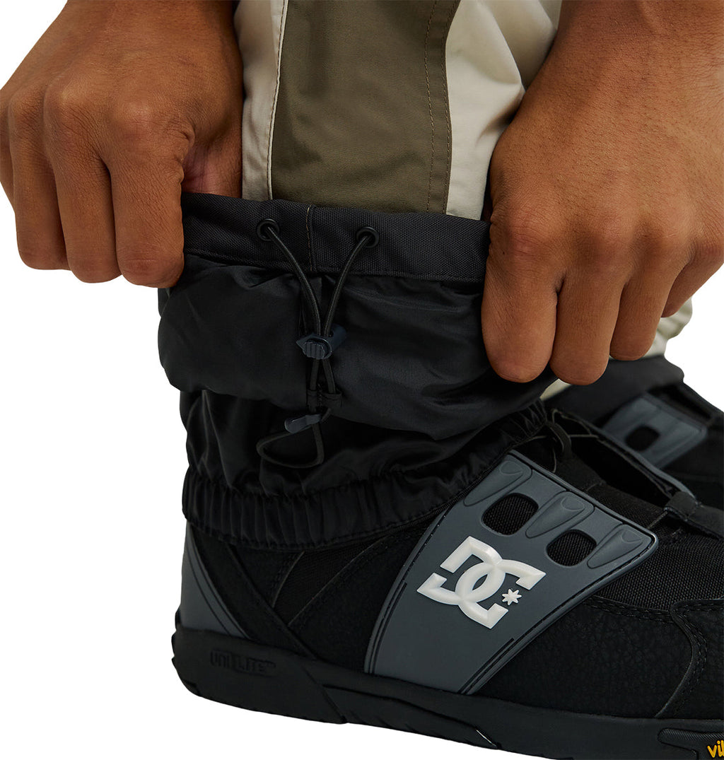 Close-up of hands adjusting black Mountain Lab Bib 20K snow gaiters by DC Shoes, designed for breathable protection, over tan pants and black DC snowboarding boots.